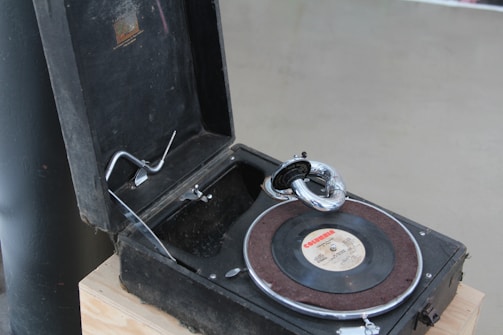 A vintage gramophone with an open lid, displaying a black vinyl record on the platter. The gramophone features a metal tonearm and a classic horn style speaker. The case and interior of the gramophone appear to be worn and dusty, indicating its age.