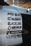A close-up view of a car's body with several automotive brand logos displayed. The logos include various companies like Blitz, Cusco, Blackline, Verus Engineering, and Michelin, along with the Toyota logo and some Japanese text.