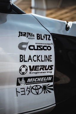 A close-up view of a car's body with several automotive brand logos displayed. The logos include various companies like Blitz, Cusco, Blackline, Verus Engineering, and Michelin, along with the Toyota logo and some Japanese text.