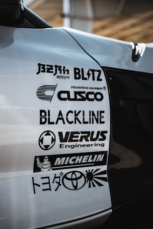 A close-up view of a car's body with several automotive brand logos displayed. The logos include various companies like Blitz, Cusco, Blackline, Verus Engineering, and Michelin, along with the Toyota logo and some Japanese text.