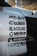 A close-up view of a car's body with several automotive brand logos displayed. The logos include various companies like Blitz, Cusco, Blackline, Verus Engineering, and Michelin, along with the Toyota logo and some Japanese text.