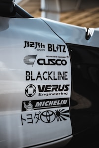A close-up view of a car's body with several automotive brand logos displayed. The logos include various companies like Blitz, Cusco, Blackline, Verus Engineering, and Michelin, along with the Toyota logo and some Japanese text.