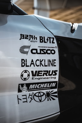 A close-up view of a car's body with several automotive brand logos displayed. The logos include various companies like Blitz, Cusco, Blackline, Verus Engineering, and Michelin, along with the Toyota logo and some Japanese text.