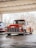 A classic red pickup truck is parked in a covered area with a backdrop of bare trees. The truck has a shiny, well-maintained exterior with prominent front grille and round headlights. The interior appears dark through the windows, and the vehicle is positioned on a smooth concrete surface under a metal roof.