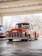 A classic red pickup truck is parked in a covered area with a backdrop of bare trees. The truck has a shiny, well-maintained exterior with prominent front grille and round headlights. The interior appears dark through the windows, and the vehicle is positioned on a smooth concrete surface under a metal roof.