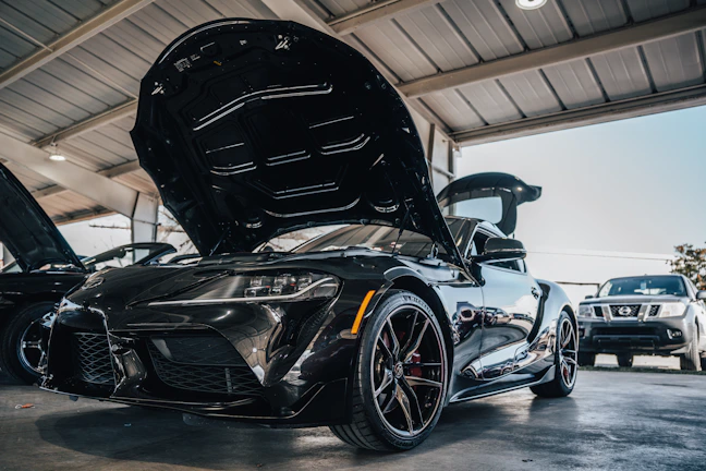 Close-up of a sleek supercar's engine bay with polished metal and carbon fiber details under bright garage lighting.
