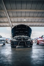 A black sports car with its hood open is parked inside a garage with other vehicles. The garage has a structured metallic roof, and there is a light fixture illuminating the area. Several other cars are parked alongside the featured car, including a red convertible and a white car, creating a showroom or service area atmosphere.