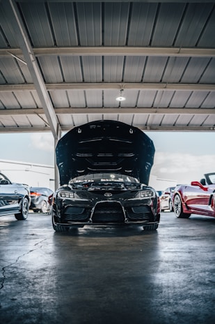 A black sports car with its hood open is parked inside a garage with other vehicles. The garage has a structured metallic roof, and there is a light fixture illuminating the area. Several other cars are parked alongside the featured car, including a red convertible and a white car, creating a showroom or service area atmosphere.