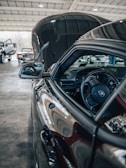 A close-up of the inside of a car with the focus on the steering wheel and side mirror. The hood is open, showing a reflection on its polished surface. In the background, there are other vehicles parked inside a large garage with a high ceiling.