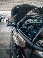 A close-up of the inside of a car with the focus on the steering wheel and side mirror. The hood is open, showing a reflection on its polished surface. In the background, there are other vehicles parked inside a large garage with a high ceiling.
