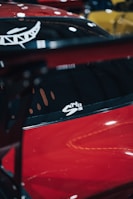 A red car with a glossy finish, featuring a white decal on the window. The background includes blurred shapes in various colors, suggesting the scene might be a car showroom or event.