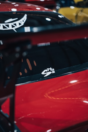 A red car with a glossy finish, featuring a white decal on the window. The background includes blurred shapes in various colors, suggesting the scene might be a car showroom or event.