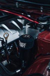 Close-up photo of a car engine featuring a black cylindrical component with a soft cover displaying the words 'CHASE BAYS' in white text. Various metallic engine parts, including hoses and a dipstick, are visible, with predominant colors of red, black, and silver. The setting appears industrial, highlighting the engineered details.