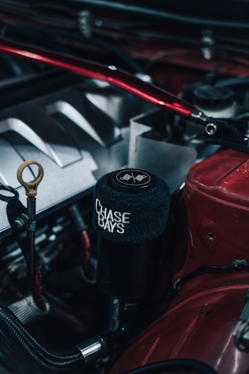 Close-up photo of a car engine featuring a black cylindrical component with a soft cover displaying the words 'CHASE BAYS' in white text. Various metallic engine parts, including hoses and a dipstick, are visible, with predominant colors of red, black, and silver. The setting appears industrial, highlighting the engineered details.