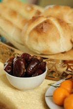A white bowl filled with dark brown dates is placed in the foreground. Behind it, a decorative golden tray holds two round loaves of freshly baked bread with a light brown crust. To the right, a white plate with small orange fruits resting on a gold tablecloth can be seen.