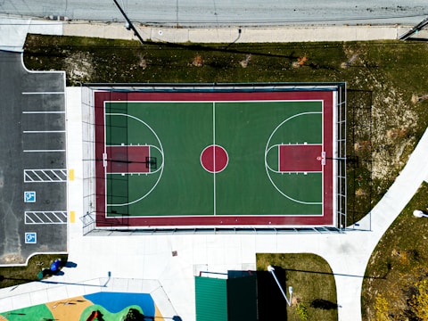 An aerial view of a basketball court located in an outdoor setting. The court's surface is painted green with maroon borders and white lines marking the playing areas. Adjacent to the court is a gray parking lot with a few handicap parking spaces marked in blue and white. Surrounding the court are patches of green grass and concrete pathways. A small playground area with colorful surfaces is visible near the bottom of the image.
