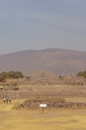 A large ancient stone pyramid is situated in the center of a vast archaeological site. Surrounding the pyramid, there are scattered smaller stone structures and platforms. A few people are walking and exploring the area. The sky is clear with a light blue hue, and the backdrop features rolling hills and sparse vegetation.