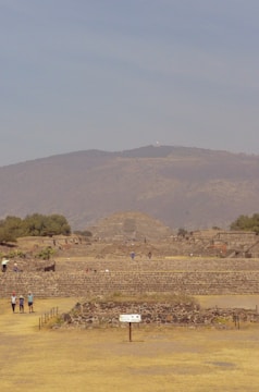 A large ancient stone pyramid is situated in the center of a vast archaeological site. Surrounding the pyramid, there are scattered smaller stone structures and platforms. A few people are walking and exploring the area. The sky is clear with a light blue hue, and the backdrop features rolling hills and sparse vegetation.