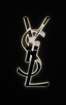 A stylized metallic monogram consisting of overlapping letters Y, S, and L is displayed against a dark background. The letters have a shiny, reflective surface and are illuminated with a subtle halo effect.