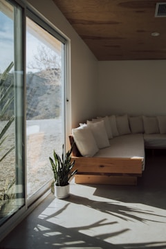 A cozy, minimalistic living space with a wooden ceiling and floor featuring a light-colored sectional sofa adorned with several soft cushions. Natural light pours in through a large glass window, casting clear shadows on the floor. A potted plant adds a touch of greenery to the room.