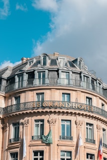 A grand, ornate hotel building with classic architectural features, including decorative ironwork balconies and a mansard roof. The front facade features numerous windows and flags, with a clear blue sky and some fluffy clouds in the background.