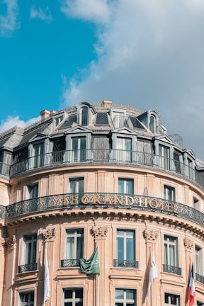 A grand, ornate hotel building with classic architectural features, including decorative ironwork balconies and a mansard roof. The front facade features numerous windows and flags, with a clear blue sky and some fluffy clouds in the background.