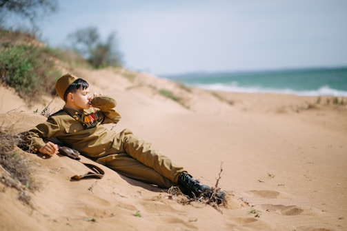 A young person dressed in a military uniform is reclining on a sandy beach, drinking from a canteen. Medals are pinned to their chest. The ocean waves are visible in the background, and the area is surrounded by sparse vegetation.