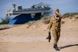 A person dressed in a military uniform with medals is walking on a sandy beach, holding a black object in one hand and shielding their eyes with the other. In the background, there is a large, elevated ship model mounted on a blue structure, and some sparse vegetation.