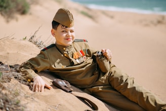 A young person in a military uniform with various medals rests on sandy terrain, holding a canteen. The background features a beach-like setting with visible water and sparse vegetation.