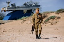 A person dressed in military uniform, adorned with medals, is walking on a sandy area. In the background, there is an old ship mounted as a display. The setting is outdoors with sand dunes and sparse vegetation.