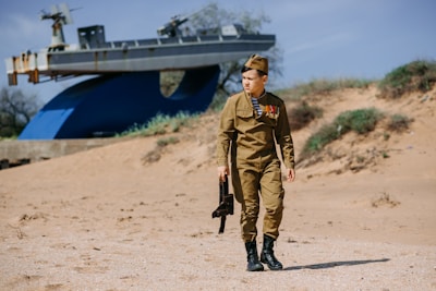 A person dressed in military uniform, adorned with medals, is walking on a sandy area. In the background, there is an old ship mounted as a display. The setting is outdoors with sand dunes and sparse vegetation.