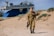 A person dressed in military uniform, adorned with medals, is walking on a sandy area. In the background, there is an old ship mounted as a display. The setting is outdoors with sand dunes and sparse vegetation.