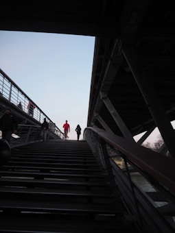 A group of people are using a modern pedestrian bridge, with one person in bright red standing out against the dark structure. The bridge has metal railings and overhead beams, with stairs leading up. The sky is clear and light, contrasting with the shadows cast by the bridge.