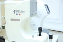 A close-up view of an ophthalmic device with digital readouts, marked with the model URK-700 and the label 'autoref keratometer.' The machine, primarily white, features a joystick for operation and various control buttons for functionality. It appears to be positioned in a clinical setting with soft lighting.