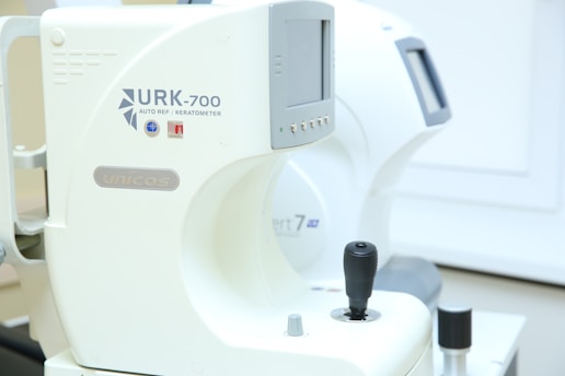 A close-up view of an ophthalmic device with digital readouts, marked with the model URK-700 and the label 'autoref keratometer.' The machine, primarily white, features a joystick for operation and various control buttons for functionality. It appears to be positioned in a clinical setting with soft lighting.