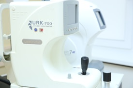 A close-up view of an ophthalmic device with digital readouts, marked with the model URK-700 and the label 'autoref keratometer.' The machine, primarily white, features a joystick for operation and various control buttons for functionality. It appears to be positioned in a clinical setting with soft lighting.