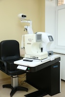 A room equipped with optical diagnostic equipment, including an autorefractor keratometer on a black adjustable table, next to a black swivel chair. The walls and floor are light-colored, and there's a window to the right.