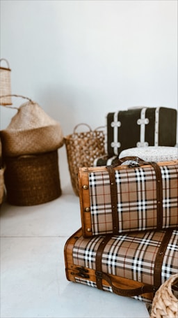 A collection of vintage suitcases and woven baskets are arranged against a plain background. The suitcases feature a distinctive plaid pattern with leather straps, while the baskets vary in texture and color, adding an earthy, rustic feel.