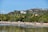 A vibrant beach scene with people enjoying the shore and shallow water. Palm trees line the sandy beach, and a lush green hillside with luxury homes overlooks the area. The sky is clear with a few distant clouds, giving a tropical, relaxed atmosphere.