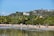 A vibrant beach scene with people enjoying the shore and shallow water. Palm trees line the sandy beach, and a lush green hillside with luxury homes overlooks the area. The sky is clear with a few distant clouds, giving a tropical, relaxed atmosphere.