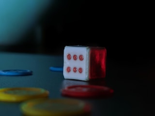 A white die with red dots sits on a surface, with the number six facing the viewer. Surrounding the die are several poker chips in blue, yellow, and red. The background is dark, creating a dramatic contrast with the brightly colored objects.