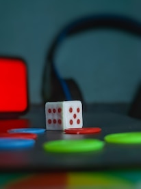 A white die with red dots is positioned on a surface surrounded by colorful circular tokens in red, blue, and green. In the background, there is a blurred set of headphones and a red rectangular object, creating a playful and colorful atmosphere.