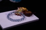 A bracelet made of transparent blue beads is placed on a textured white surface alongside dried flowers with hues of yellow and brown. The background is a deep black, adding contrast to the composition.