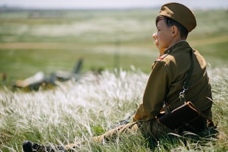A person wearing a military-style uniform sits on a grassy hillside. They are looking into the distance, with one hand raised to their face. In the background, an indistinct aircraft shape is visible, suggesting a possible connection to a historical or military setting.