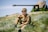 A young person wearing a military-style uniform sits thoughtfully on a grassy field. Medals are pinned to the uniform, and a vintage artillery piece is out of focus in the background, suggesting a historical or military setting. The greenery is lush, with sunlight casting gentle shadows.