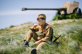A young person wearing a military-style uniform sits thoughtfully on a grassy field. Medals are pinned to the uniform, and a vintage artillery piece is out of focus in the background, suggesting a historical or military setting. The greenery is lush, with sunlight casting gentle shadows.