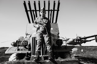 A young person in military uniform sits on the hood of an old military vehicle with several rocket launchers mounted on the back. In the background, there are two vintage airplanes, one of which is partially visible. The image is in black and white and conveys a historical or wartime scene.