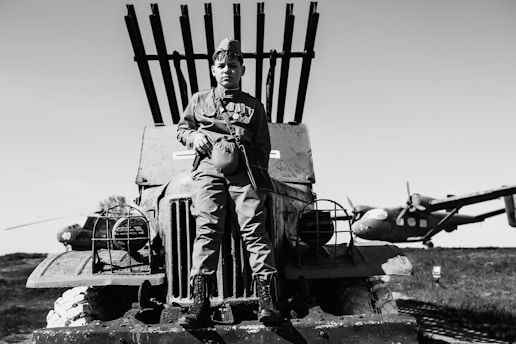 A young person in military uniform sits on the hood of an old military vehicle with several rocket launchers mounted on the back. In the background, there are two vintage airplanes, one of which is partially visible. The image is in black and white and conveys a historical or wartime scene.