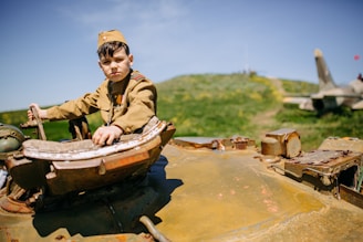 A young boy wearing a military-style uniform is seated on top of an old, rusted tank. The background shows a green hill and a cloudy sky, with a jet plane visible in the distance, suggesting a military or war-themed environment. The boy looks towards the camera, and the landscape is bright and sunny.