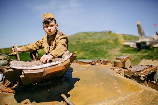 A young boy wearing a military-style uniform is seated on top of an old, rusted tank. The background shows a green hill and a cloudy sky, with a jet plane visible in the distance, suggesting a military or war-themed environment. The boy looks towards the camera, and the landscape is bright and sunny.